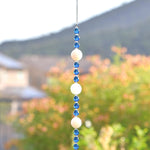 Long sun catcher from Cinjo Handmade hanging from a hook in front of a foliage background. Sun catcher has a 4 inch loop at the top and includes 3 large round beads made of white quartz. Two of these quartz beads are more off white or cream colored. There are 3 smaller deep aqua blue round glass beads between each of the quartz beads. A round glass crystal prism anchors the piece.