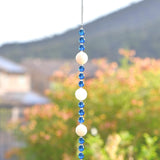 Long sun catcher from Cinjo Handmade hanging from a hook in front of a foliage background. Sun catcher has a 4 inch loop at the top and includes 3 large round beads made of white quartz. Two of these quartz beads are more off white or cream colored. There are 3 smaller deep aqua blue round glass beads between each of the quartz beads. A round glass crystal prism anchors the piece.