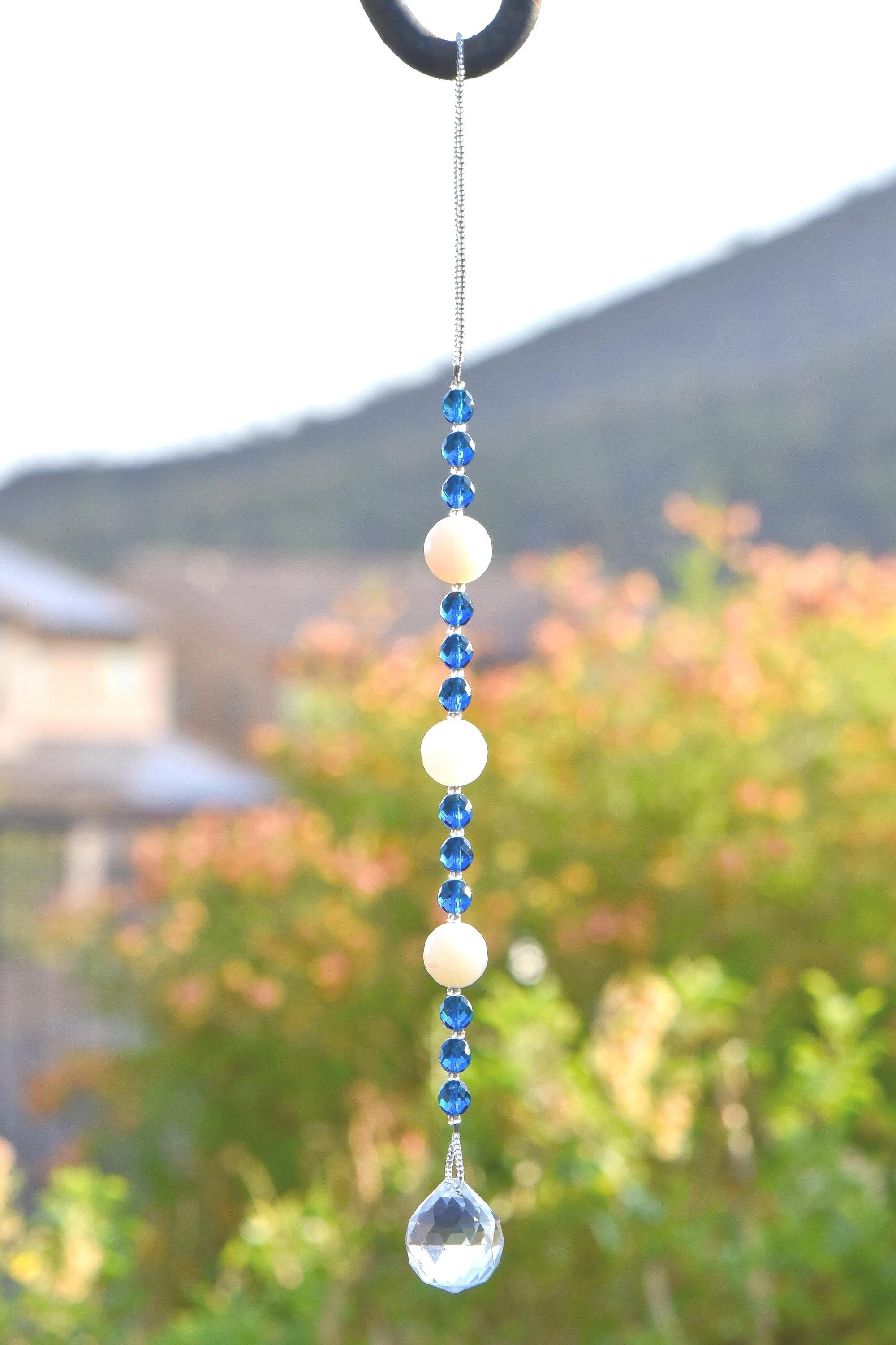 Long sun catcher from Cinjo Handmade hanging from a hook in front of a foliage background. Sun catcher has a 4 inch loop at the top and includes 3 large round beads made of white quartz. Two of these quartz beads are more off white or cream colored. There are 3 smaller deep aqua blue round glass beads between each of the quartz beads. A round glass crystal prism anchors the piece.