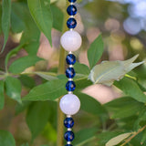 Close view of the middle of a Cinjo Handmade sun catcher hanging from a tree. Two large round white quartz beads are shown with smaller blue glass beads in between each quartz bead.