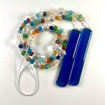 Brightly colored glass beads from Ghana, South Africa&#39;s Krobo tribe are strung with large solid clear glass beads, coiled onto a white surface. Two long rectangular pieces of deep aqua blue fused glass are at each end of the long wire.
