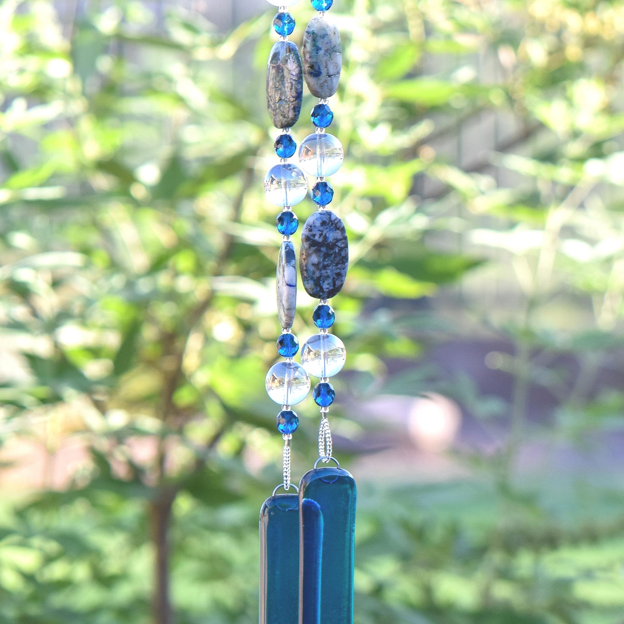 Large oval beads made of azurite and malachite gemstones are strung on wire with aqua blue glass crystal beads and large clear, solid glass beads. Two ends of a long beaded wire are hanging against a blurred landscape, anchored by two pieces of transparent aqua kiln formed glass.