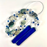 Large oval beads made of azurite and malachite gemstones are strung on wire with aqua blue glass crystal beads and large clear, solid glass beads. Two pieces of blue glass are attached at each end of the long wire. The entire piece is coiled on a white background.