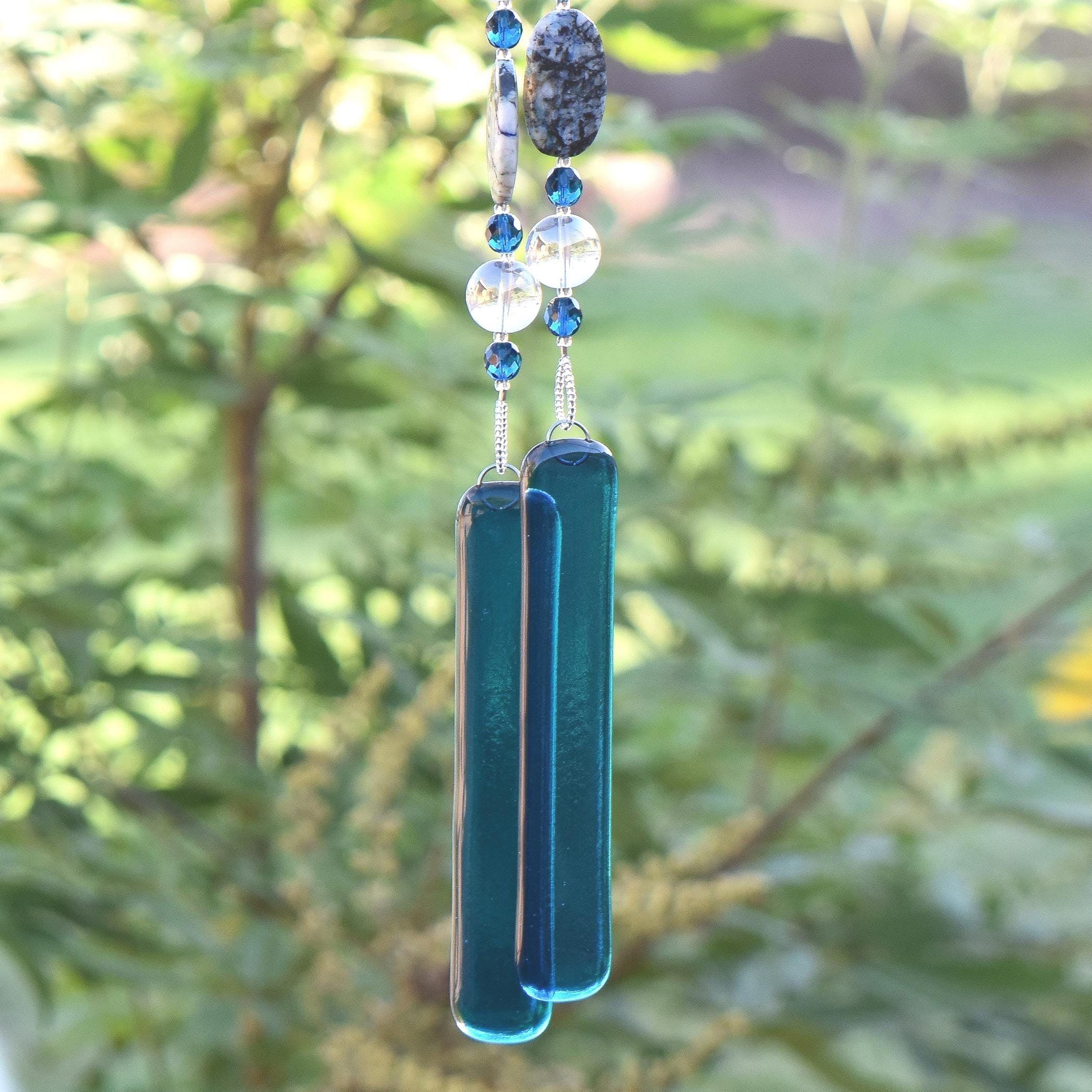 Wind chime with a series of decorative elements. The top features a dark, speckled stone and blue beads. Below, clear and blue beads are strung together, leading to two long, rectangular lue glass pieces. The chime hangs against a blurred green background.