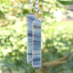 Two pieces of streaky fused glass hanging vertically from a tree. The glass has a cream background and streaks of black, gray and tan with a slight bluish/green hue. Each piece is rectangular. At the top of each glass piece are small faceted glass crystal beads.