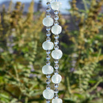Two strings of wire from a Cinjo Handmade chime with several large light green Krobo tribe beads from Ghana, Africa. Between each Krobo bead are 3 smaller, round faceted glass crystal beads. Hanging vertically against blurred background.