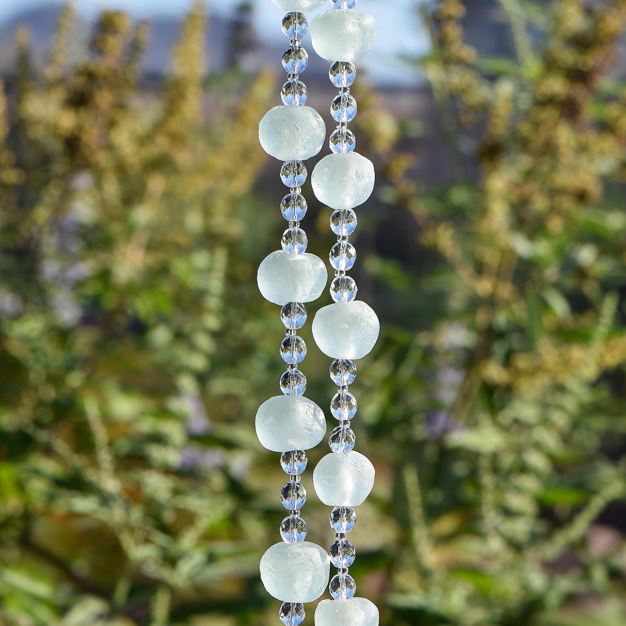 Two strings of wire from a Cinjo Handmade chime with several large light green Krobo tribe beads from Ghana, Africa. Between each Krobo bead are 3 smaller, round faceted glass crystal beads. Hanging vertically against blurred background.