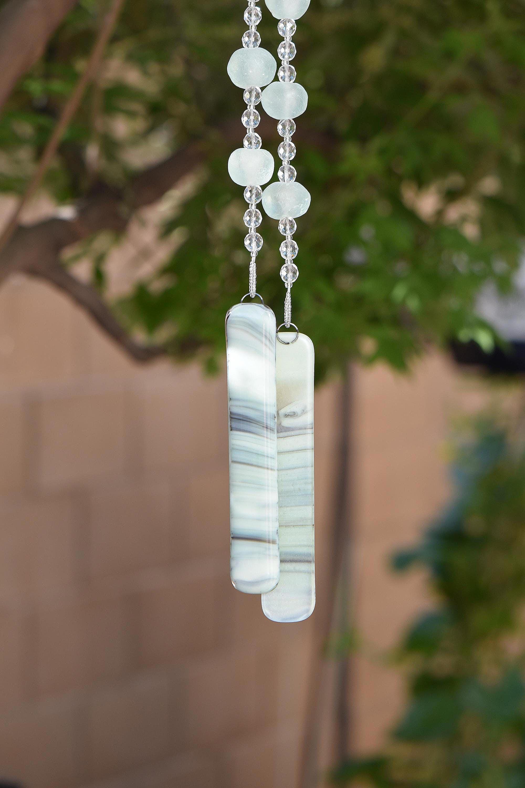 Bottom of hanging wind chime from Cinjo Handmade.Four large sea glass beads are pictured, strung with smaller clear glass beads and anchored by two pieces of kiln formed glass. The chime is hanging in front of blurred foliage.