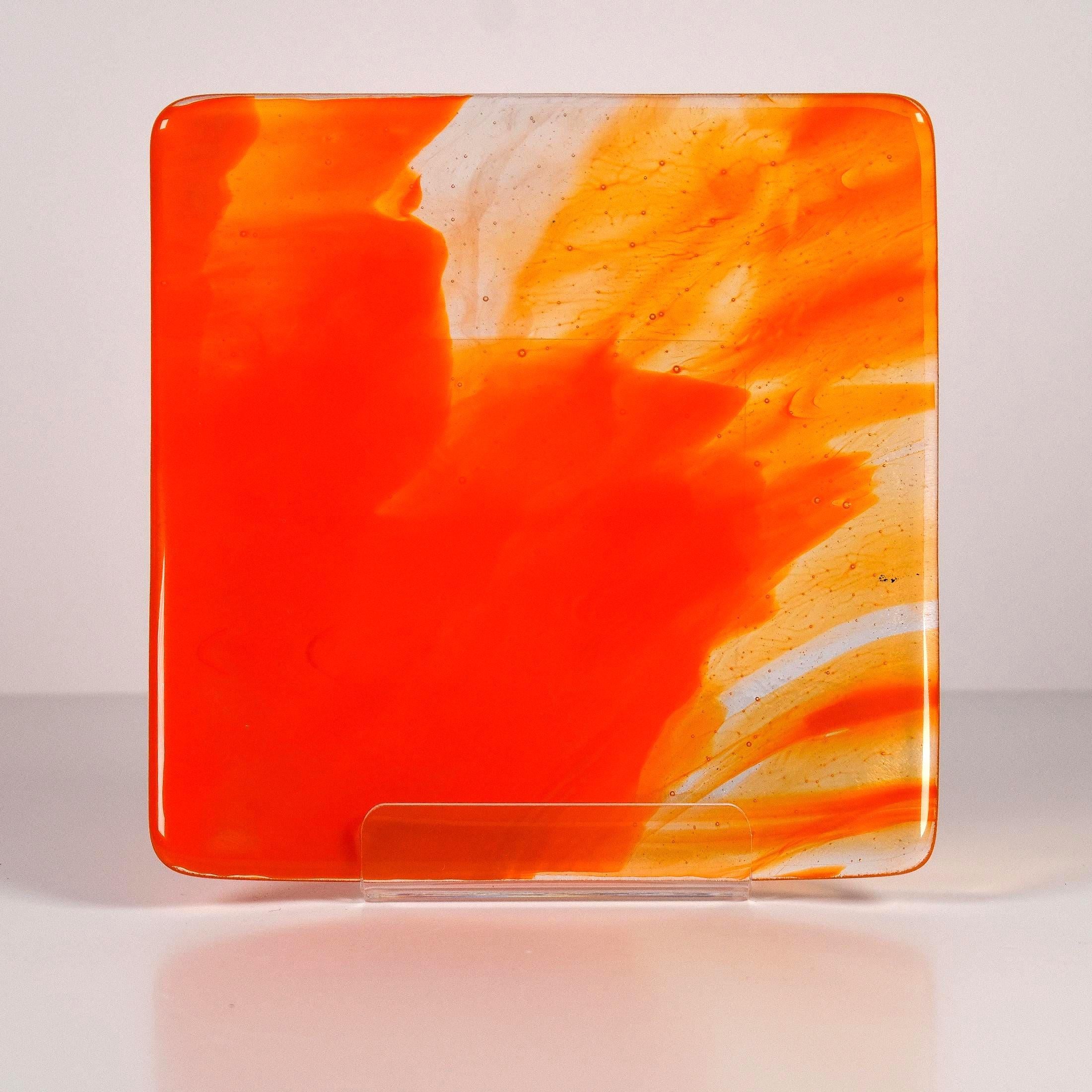 Square fused glass tray made of clear glass with streaks of bright orange, sitting on clear acrylic stand, viewed from the front. Background is white.