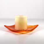 Square fused glass tray made of clear glass with streaks of bright orange holding a pillar candle. Tray is viewed from the side showing the sloped corners. Surface and background are white.