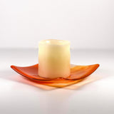 Square fused glass tray made of clear glass with streaks of bright orange holding a pillar candle. Tray is viewed from the side showing the sloped corners. Surface and background are white.