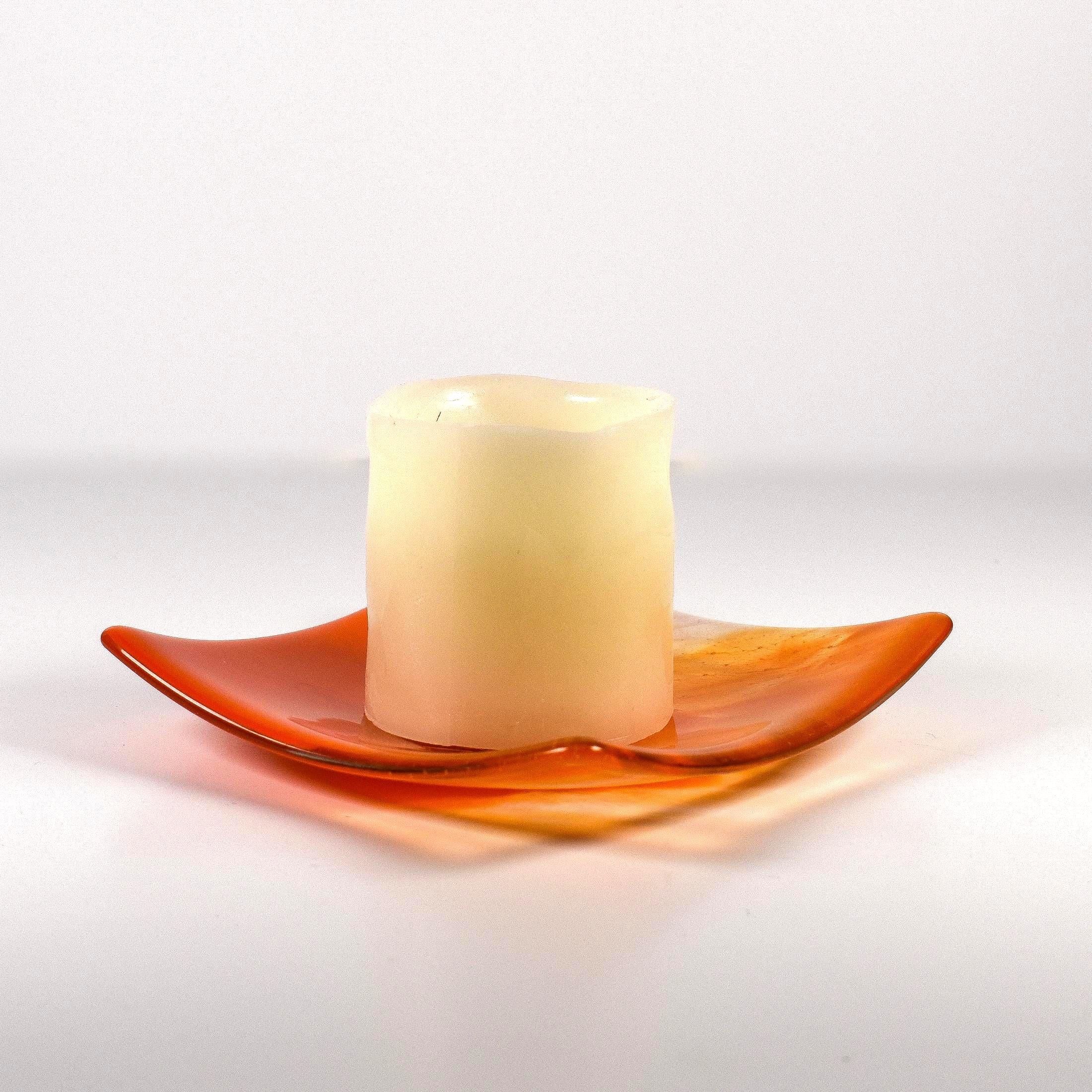 Square fused glass tray made of clear glass with streaks of bright orange holding a pillar candle. Tray is viewed from the side showing the sloped corners. Surface and background are white.