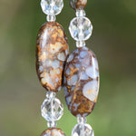 May include: Close-up of two large, oval-shaped beads fire opal stone beads with a brown and blue mottled pattern. Clear, faceted crystal beads separate the larger beads. Two strings are displayed against a blurred green background.