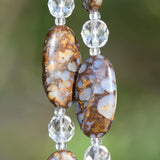 May include: Close-up of two large, oval-shaped beads fire opal stone beads with a brown and blue mottled pattern. Clear, faceted crystal beads separate the larger beads. Two strings are displayed against a blurred green background.