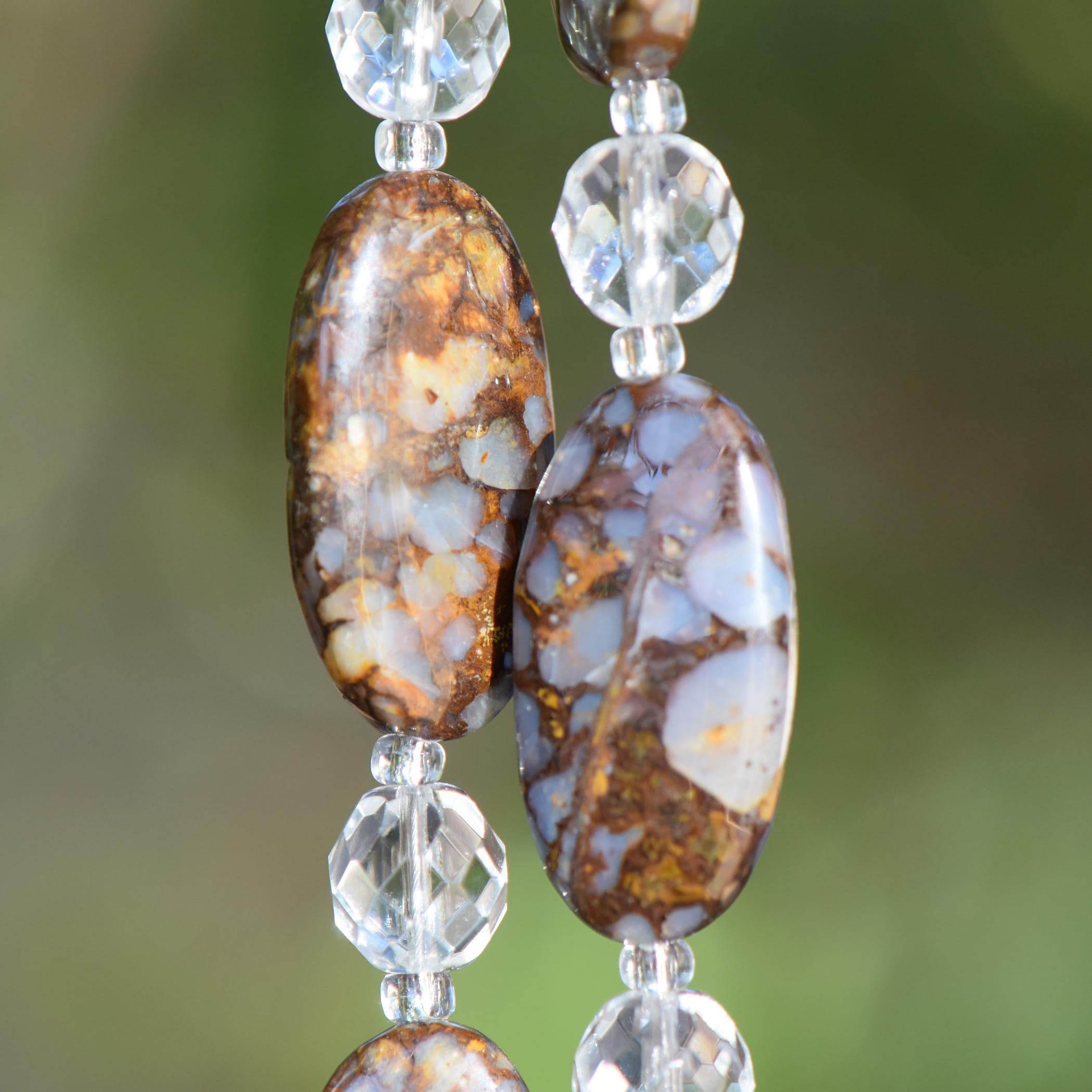 May include: Close-up of two large, oval-shaped beads fire opal stone beads with a brown and blue mottled pattern. Clear, faceted crystal beads separate the larger beads. Two strings are displayed against a blurred green background.