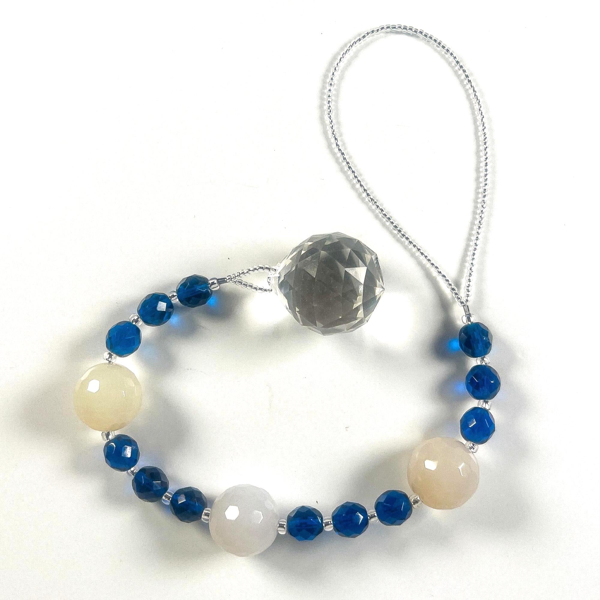Long sun catcher from Cinjo Handmade coiled on white surface. Sun catcher has a 4 inch loop at the top and includes 3 large round beads made of white quartz. Two of these quartz beads are more off white or cream colored. There are 3 smaller deep aqua blue round glass beads between each of the quartz beads. A round glass crystal prism anchors the piece.