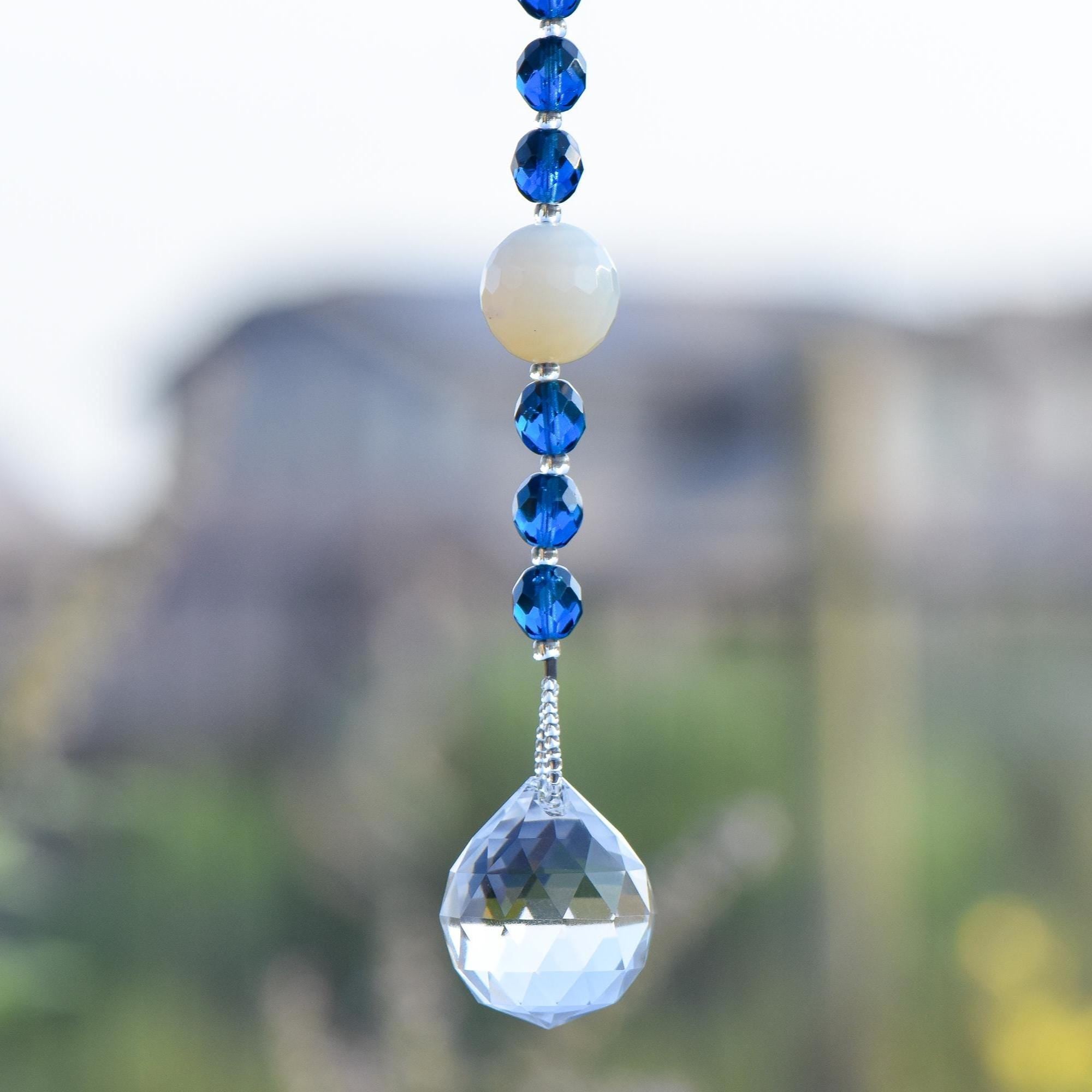 Long sun catcher from Cinjo Handmade hanging in front of blurred suburban background. One large white quartz bead is shown with 3 dark blue glass beads above and below. At the bottom is a large round glass crystal prism.