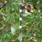 Long sun catcher from Cinjo Handmade hanging from a tree. Sun catcher has a 4 inch loop at the top and includes 3 large round beads made of white quartz. There are 3 smaller deep aqua blue round glass beads between each of the quartz beads. A round glass crystal prism anchors the piece.