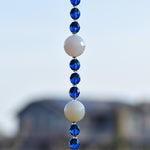 Close view of the middle of a Cinjo Handmade sun catcher hanging from a tree. Two large round white quartz beads are shown with smaller blue glass beads in between each quartz bead.