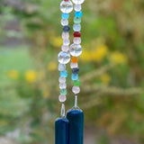 Brightly colored glass beads from Ghana, South Africa&#39;s Krobo tribe are strung with large solid clear glass beads, hanging vertically against blurred background. The two strung &quot;legs&quot; are anchored by two pieces of dark aqua blue fused glass.
