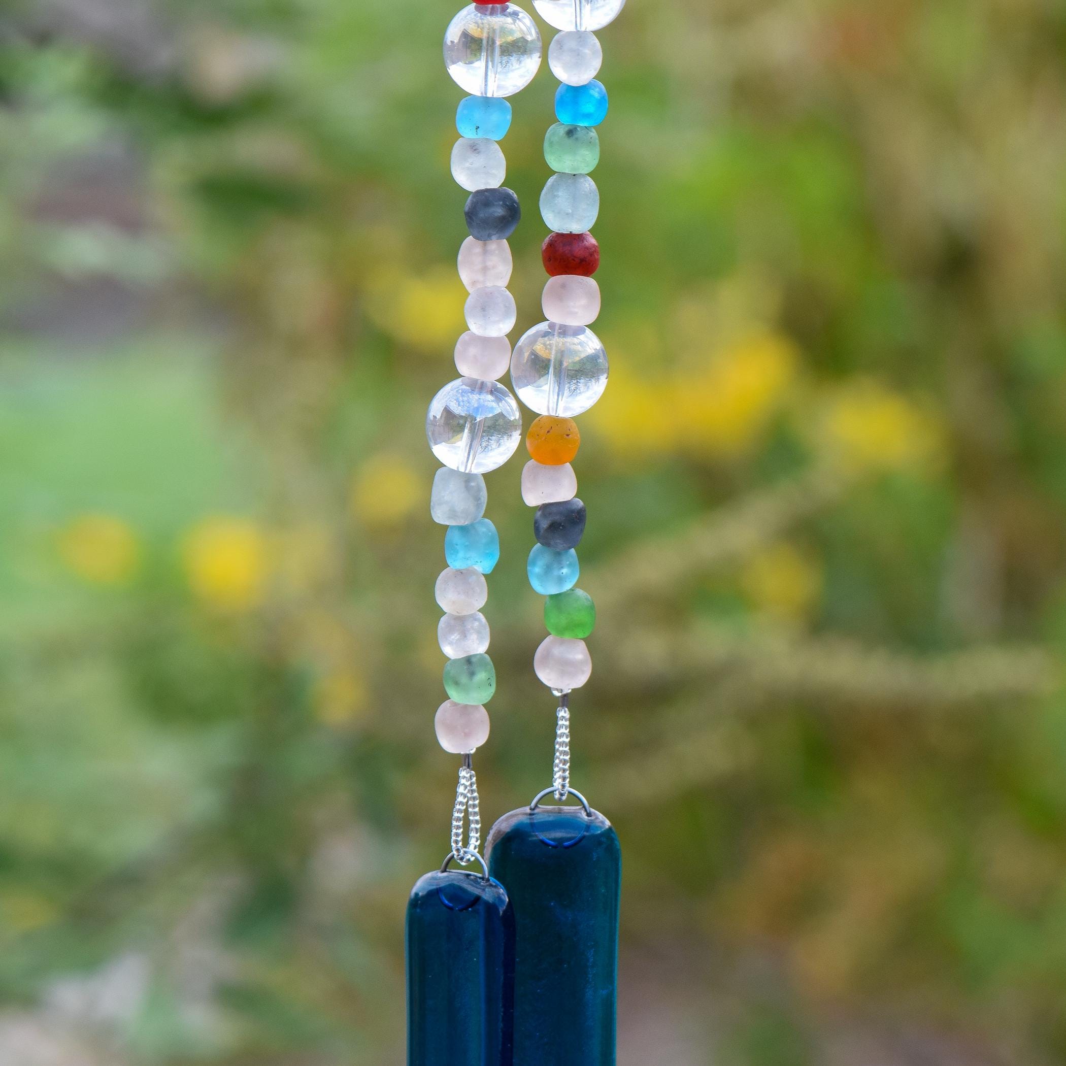 Brightly colored glass beads from Ghana, South Africa&#39;s Krobo tribe are strung with large solid clear glass beads, hanging vertically against blurred background. The two strung &quot;legs&quot; are anchored by two pieces of dark aqua blue fused glass.