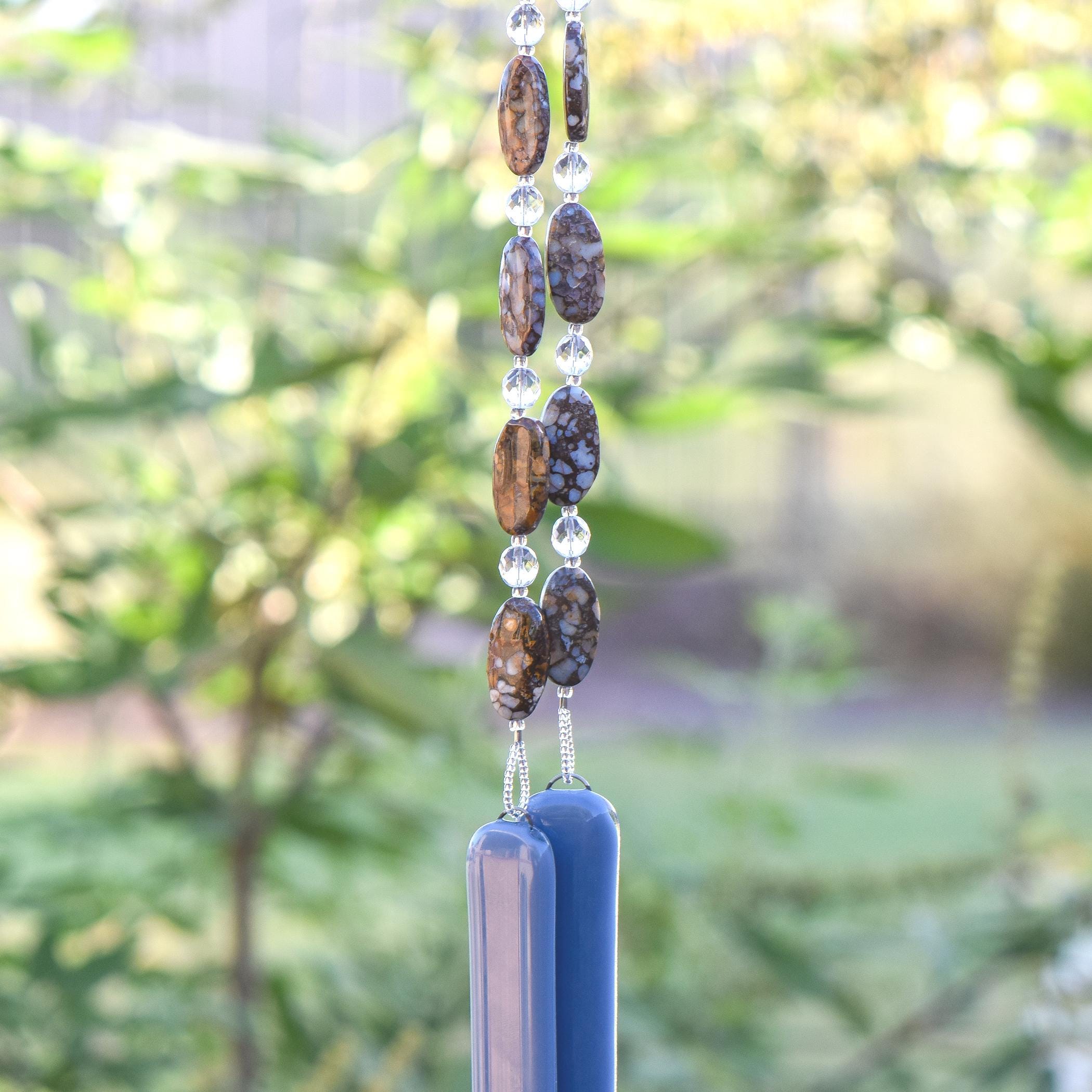 Large oval fire opal gemstone beads strung on wire with smaller clear glass beads, hanging vertically against blurred greenery background. Each of two hanging wires are anchored by rectangular pieces of grayish-blue glass.