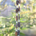 Two strands of beads with a mix of colors. The beads are a mix of oval-shaped, speckled brown and tan fire opal stones, and clear, faceted crystal beads. The beads are strung together. The background is blurred, with hints of green.