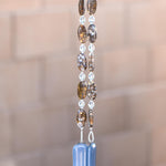 Large oval fire opal gemstone beads strung on wire with smaller clear glass beads, hanging vertically against blurred greenery background. Each of two hanging wires are anchored by rectangular pieces of grayish-blue glass.