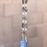 Large oval fire opal gemstone beads strung on wire with smaller clear glass beads, hanging vertically against blurred greenery background. Each of two hanging wires are anchored by rectangular pieces of grayish-blue glass.