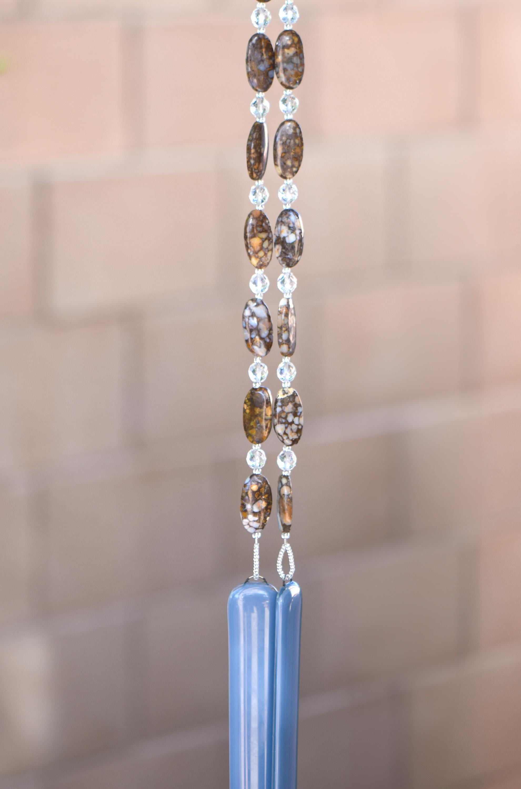 Large oval fire opal gemstone beads strung on wire with smaller clear glass beads, hanging vertically against blurred greenery background. Each of two hanging wires are anchored by rectangular pieces of grayish-blue glass.