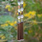 Bottom of Cinjo Handmade wind chime hanging in front of blurred foliage background. Two piece of transparent pale purple fused glass, rectangular-shaped, are anchors for two strings of beaded wire. Rhyolite stone beads are tear drop shaped and there is a larger, clear, reflective glass bead in between each stone bead. The stone beads are marked and in shades of brown, tan and gray.