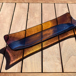 Outdoor photo on a wooden table of the fused glass channel tray, glowing amber in sunlight with deep purple ends.
