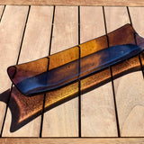 Outdoor photo on a wooden table of the fused glass channel tray, glowing amber in sunlight with deep purple ends.