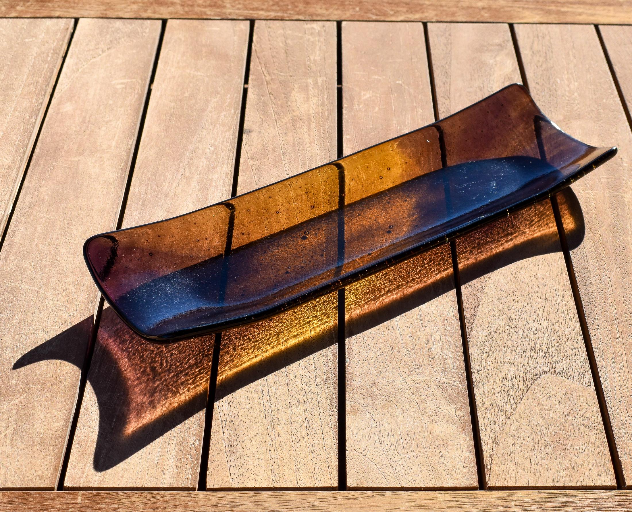 Outdoor photo on a wooden table of the fused glass channel tray, glowing amber in sunlight with deep purple ends.