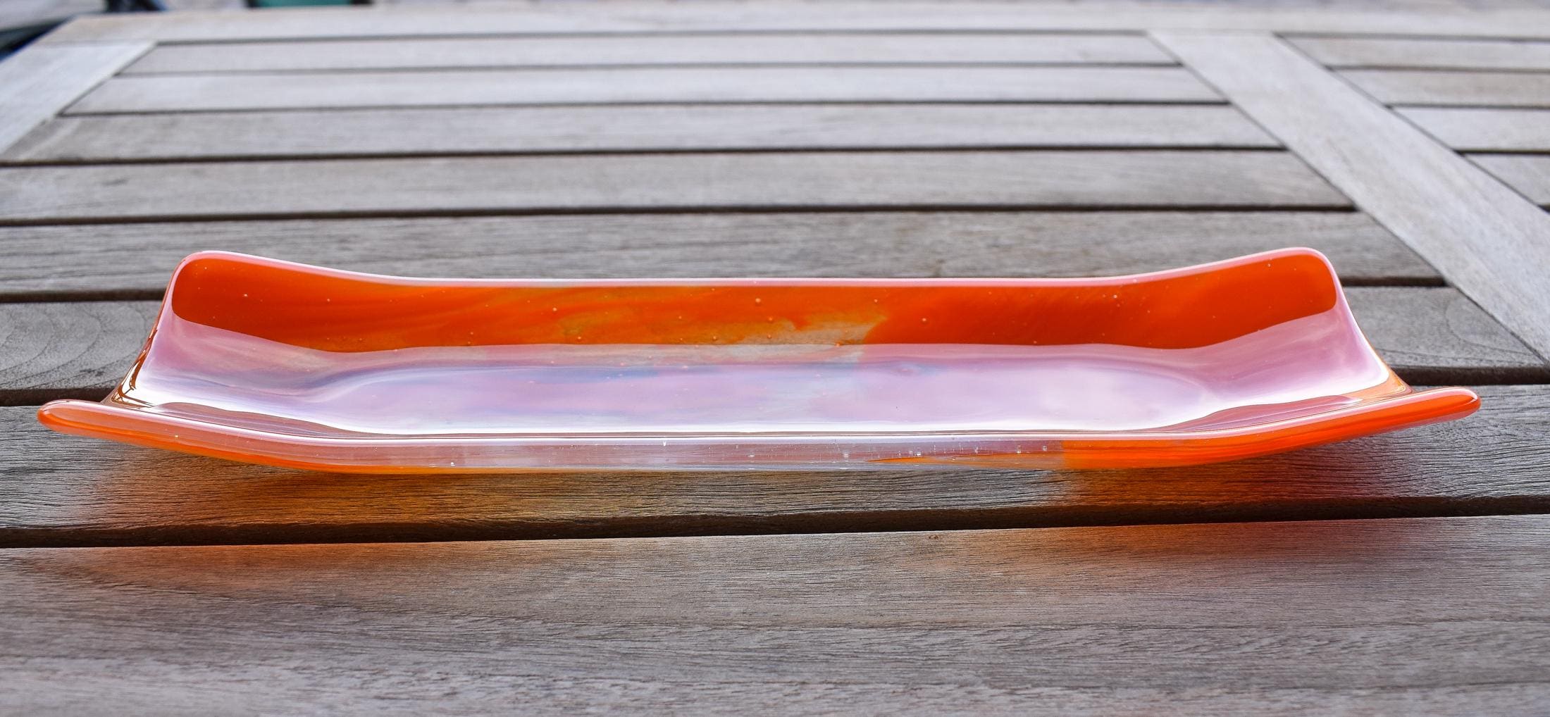 Orange and clear fused glass channel tray photographed from the side on a slatted wood table.