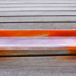 Orange and clear fused glass channel tray photographed from the side on a slatted wood table.