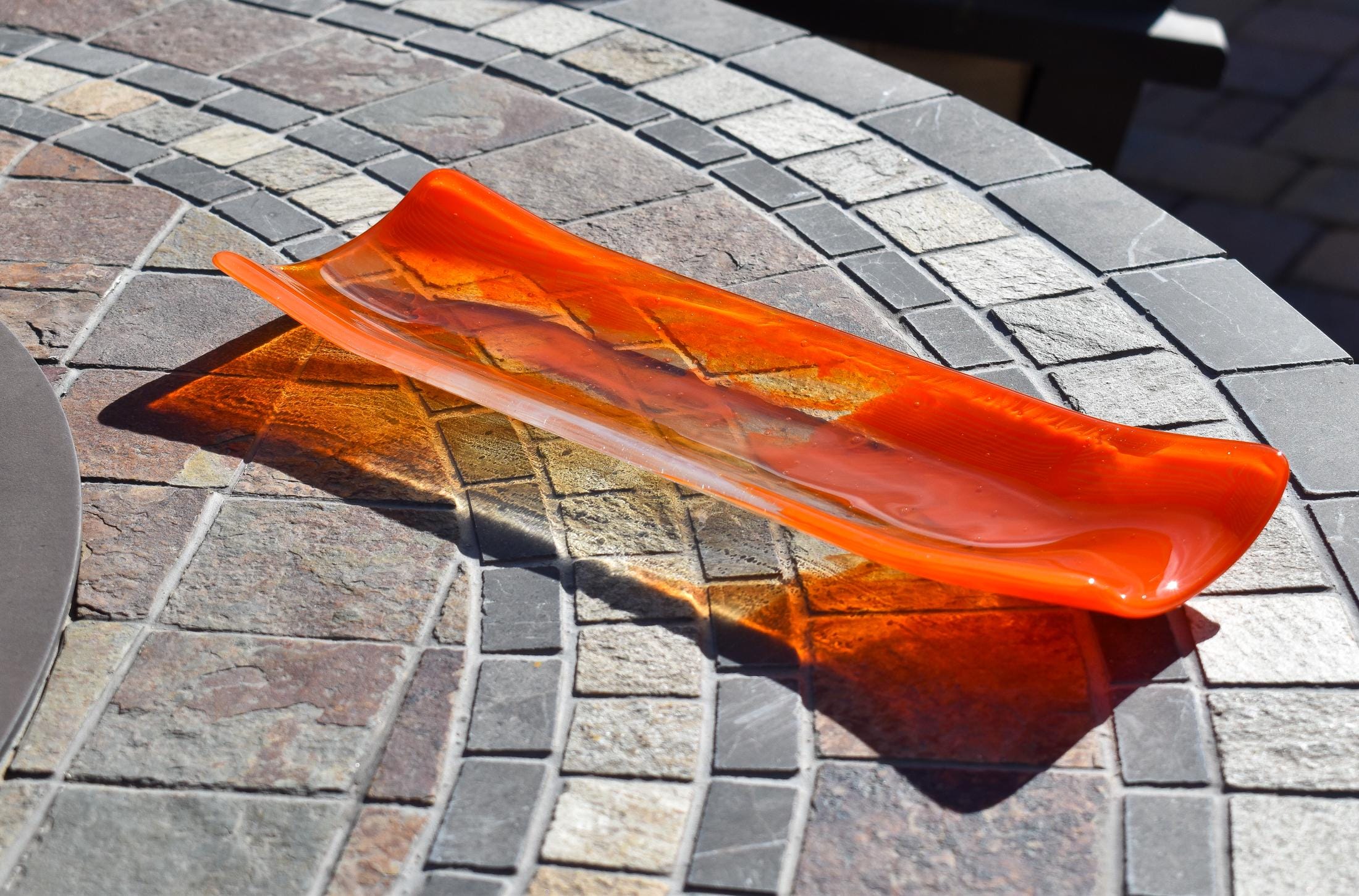 Orange and clear fused glass channel tray photographed from an angle on a stone tile table in sunlight.