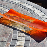 Orange and clear fused glass channel tray photographed from an angle on a stone tile table in sunlight.