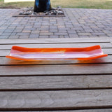 Orange and clear fused glass channel tray photographed from the side on a slatted wood table.