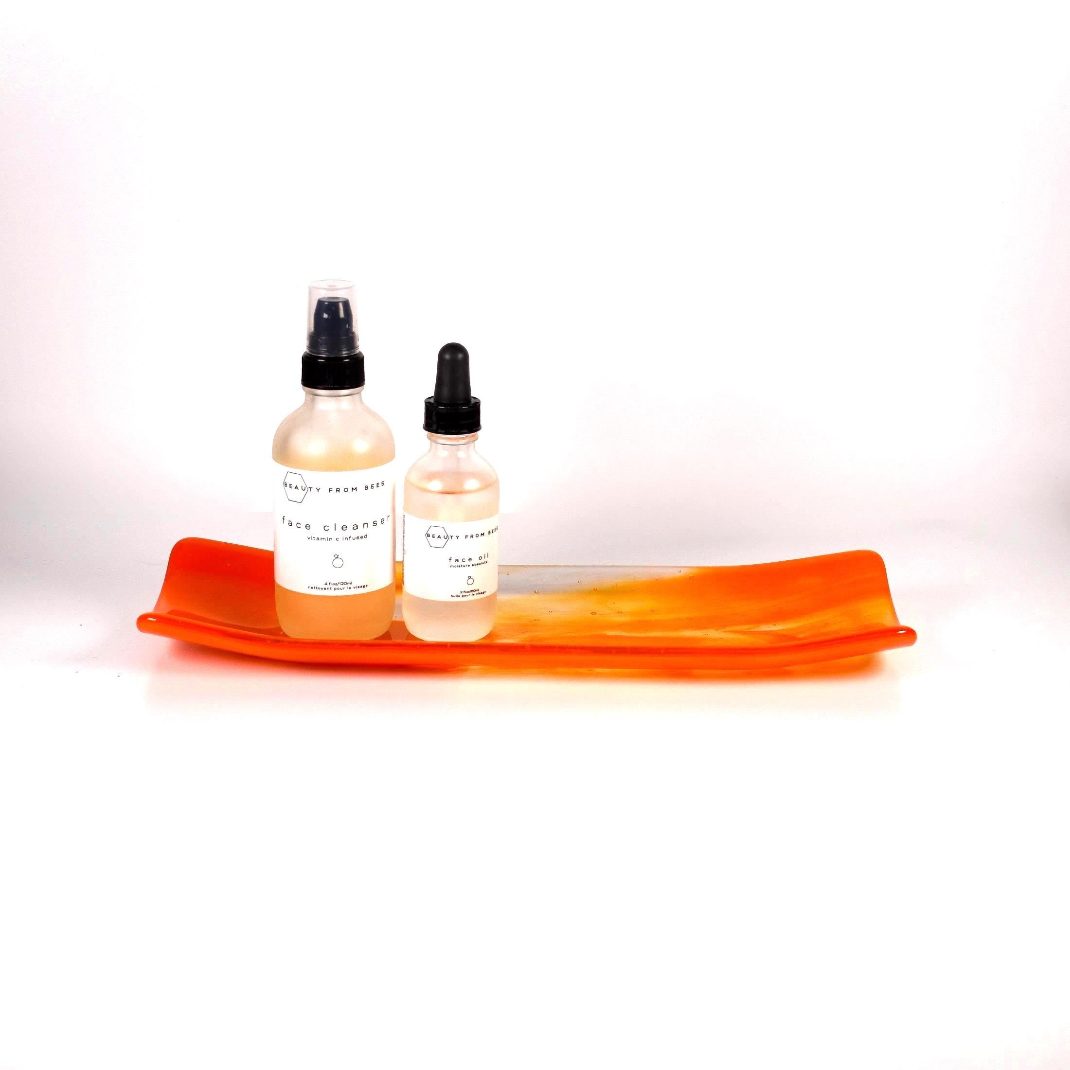 Orange and clear fused glass channel tray photographed from the side on a white background. Two bottles of bath products are on the tray.