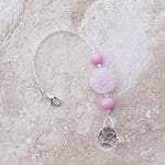 Large round carved rose quartz bead flanked by two smaller pink stone beads and anchored by round crystal prism, on marbled tile surface.