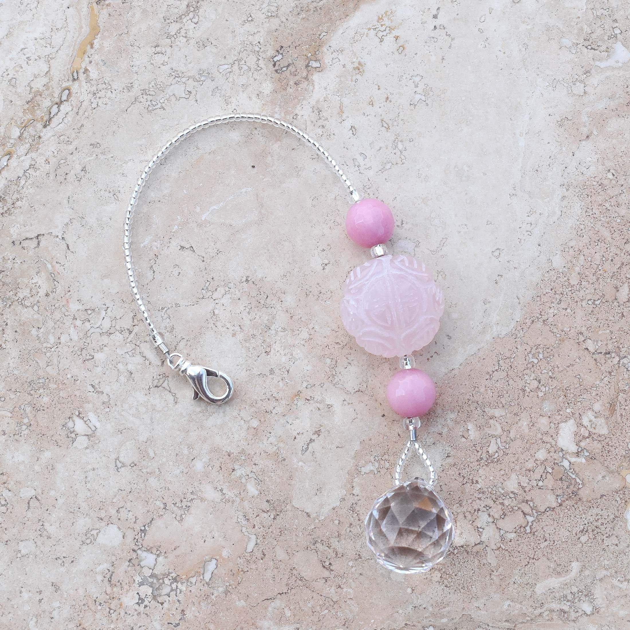 Large round carved rose quartz bead flanked by two smaller pink stone beads and anchored by round crystal prism, on marbled tile surface.