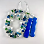 Close-up of a handmade glass wind chime featuring alternating round and frosted beads in shades of blue, green, clear, and white. The beads are strung on wire and end in two smooth cobalt blue glass pieces, all coiled on a white surface.