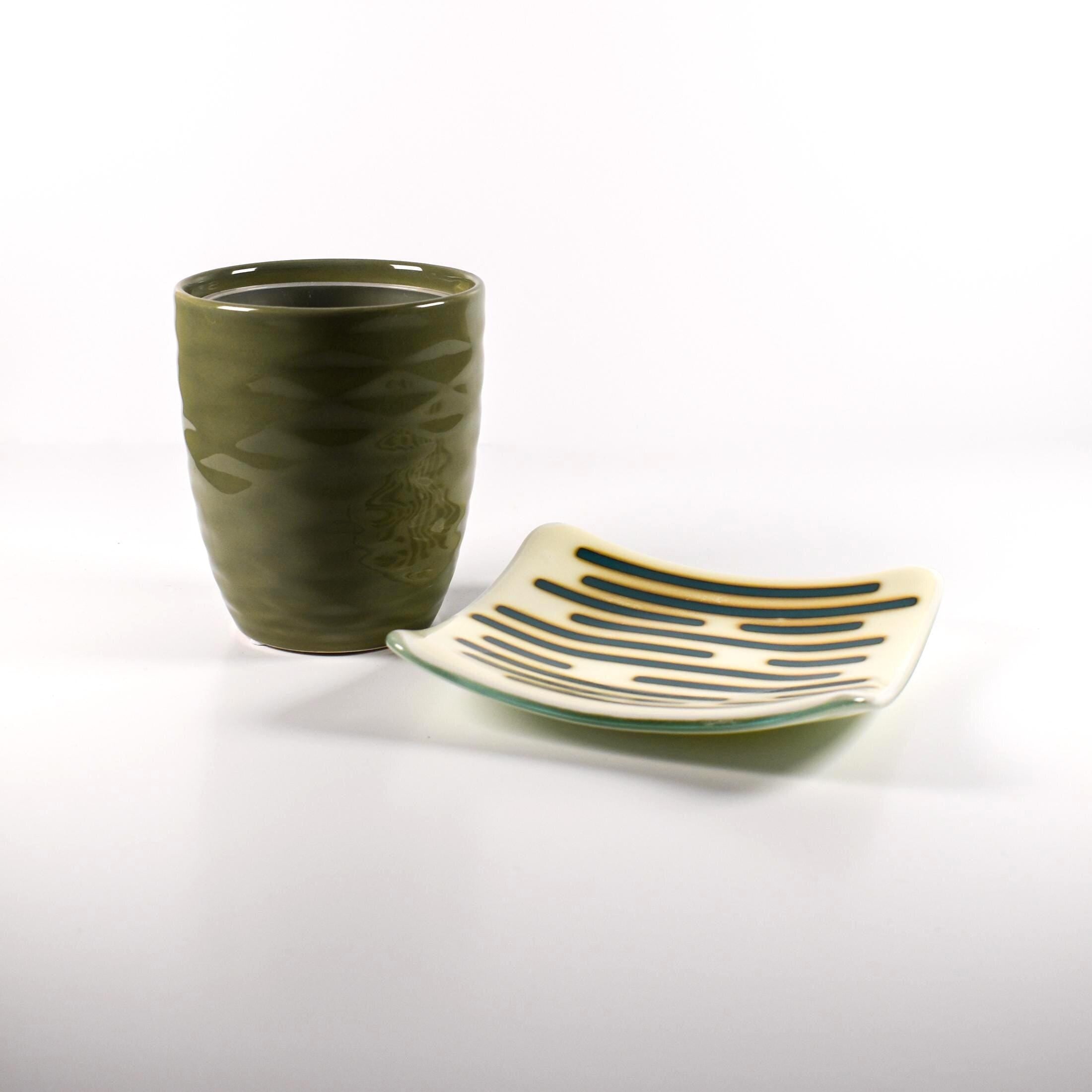 Square glass dish on a white surface with a green vase or cup next to it.  The dish has a base color of vanilla cream with abstract design consisting of lines of peacock green.