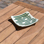 Square glass dish, viewed from above at an angle, on a slatted wood table. The dish has a base color of vanilla cream with abstract design consisting of lines of peacock green.