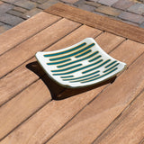 Square glass dish, viewed from above at an angle, on a slatted wood table. The dish has a base color of vanilla cream with abstract design consisting of lines of peacock green.