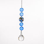 Small sun catcher hanging vertically against white background. On the stainless steel string are 4 medium blue glass beads in the Murano glass style and one larger Murano glass bead that is blue with gold and silver foil. A round glass crystal prism provides the anchor.