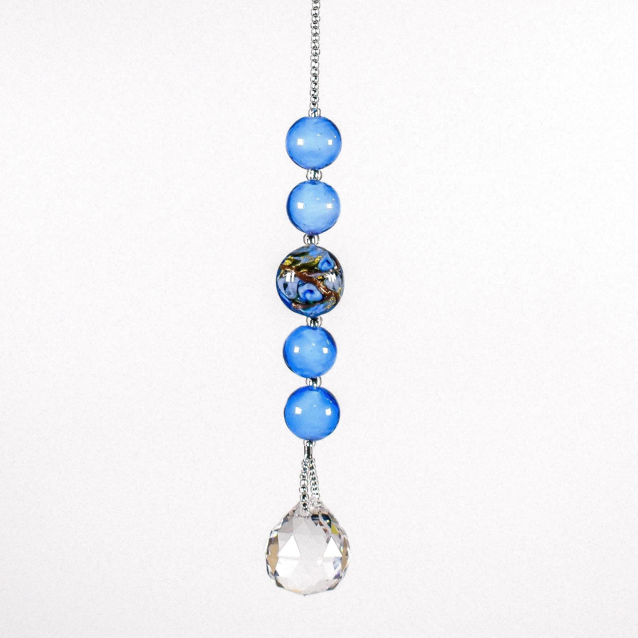Small sun catcher hanging vertically against white background. On the stainless steel string are 4 medium blue glass beads in the Murano glass style and one larger Murano glass bead that is blue with gold and silver foil. A round glass crystal prism provides the anchor.