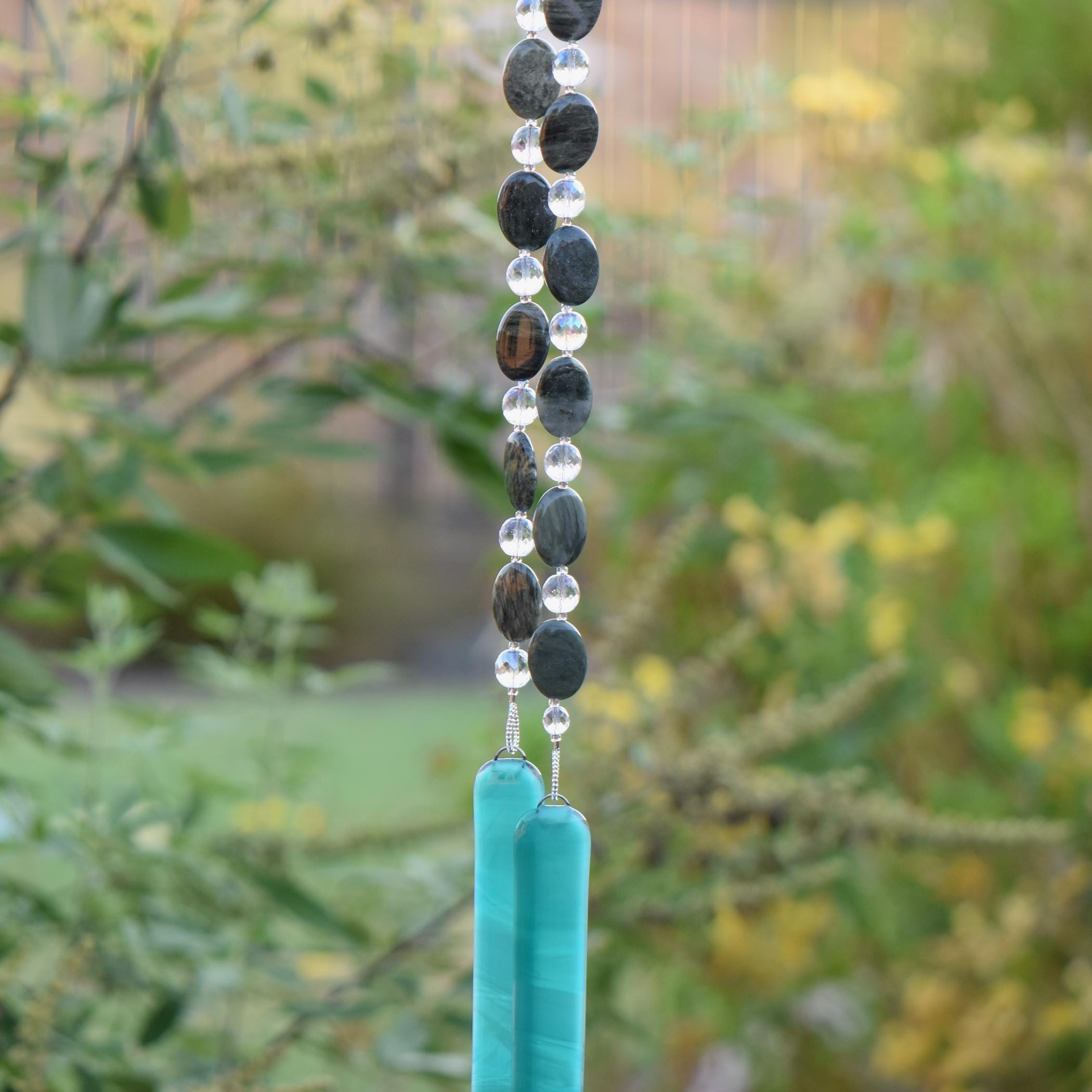 Large gray-green oval stone beads strung with clear glass faceted and reflective beads, hanging in two strands vertically; anchored by two pieces of teal and white fused glass, against blurred landscape background.