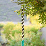 Large gray-green oval stone beads strung with clear glass lamp worked beads, hanging in two strands vertically; anchored by two pieces of teal and white fused glass, against blurred landscape background.